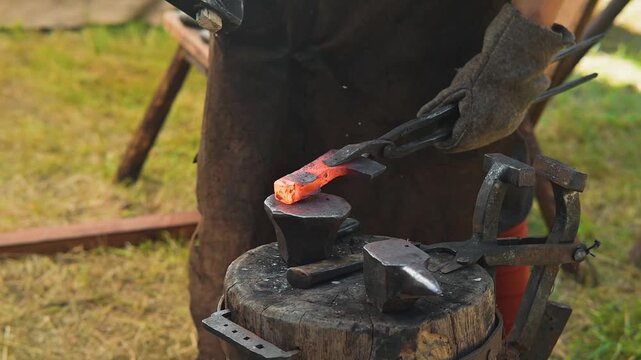 A blacksmith shapes a piece of red-hot metal using tongs and a hammer. The activity takes place outdoors in a workshop with simple tools and equipment