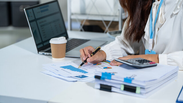 Businesswoman working at office desk with laptop, calculator and printed charts, checking financial data and planning budget during the workday.