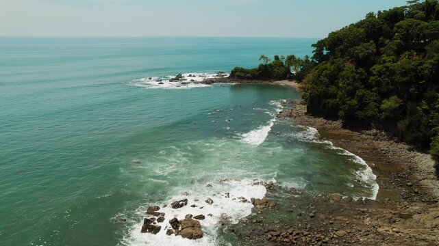 Drone view of Pacific Ocean waves crash on rocky tropical cove below green forest coastline