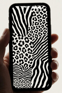 Person holds smartphone showing abstract animal print collage on screen. Black, white zebra stripes, leopard spots pattern display on mobile device. Close up of hand using phone with modern wild