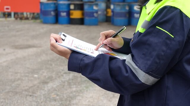 Checking on chemical hazardous safety checklist form, close-up at worker hand, with factory warehouse and metal barrel storage as background. Industrial safe work practice scene, selective focus.