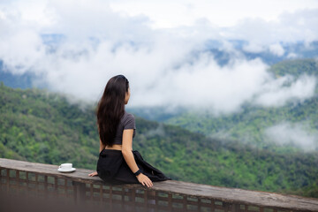 Fototapeta premium Woman sitting on wooden balcony overlooking misty mountain valley, enjoying peaceful solitude among lush green hills and drifting cloud
