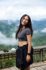 Fototapeta premium Young woman in dark shirt standing on wooden terrace above misty mountain valley, eyes closed with peaceful expression and wind moving her hair