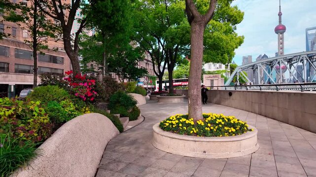 A curved riverside paved walkway lined with trees and flower beds, with distant urban skyline and steel bridge under clear sky.