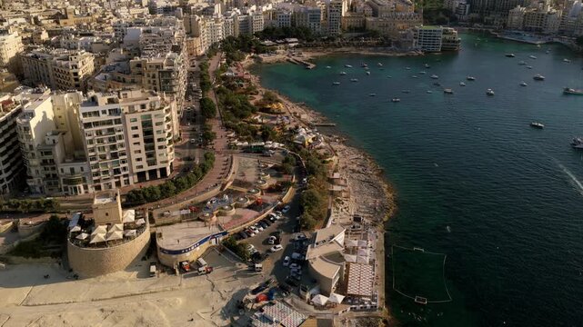 Drone flies along Malta bayfront with apartments, rocky shoreline, parked cars and boats in calm water.