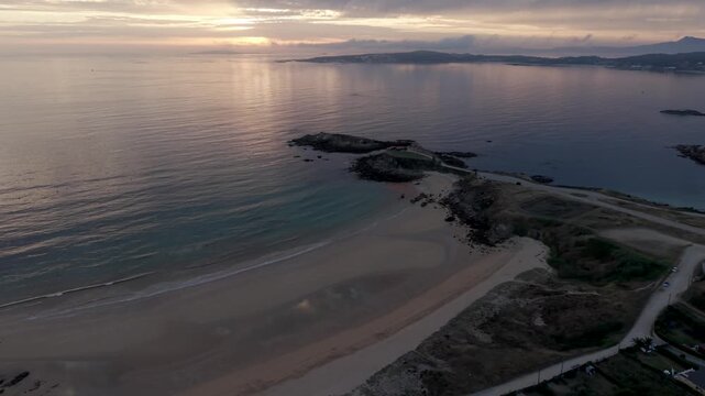 Aerial drone approach toward A Lanzada beach and its rocky headland at sunset with soft golden light on the calm sea in Galicia Spain