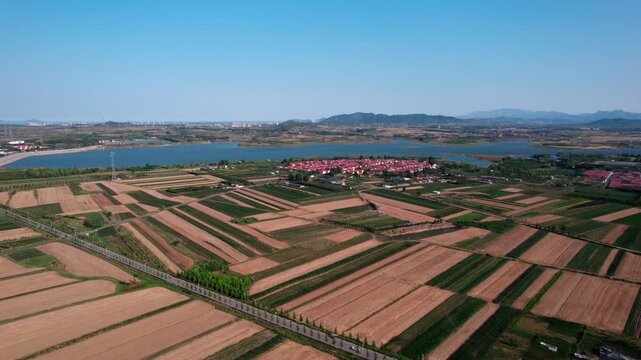 Aerial landscape showing patchwork farmland, a lake, and a village under a clear blue sky with distant hills.