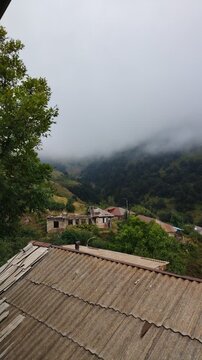 Thick fog and clouds rolling over a mountain village and forest. Beautiful view of the mountains in the morning. Fog covered the houses of the village. Green forests and fields and white fog.