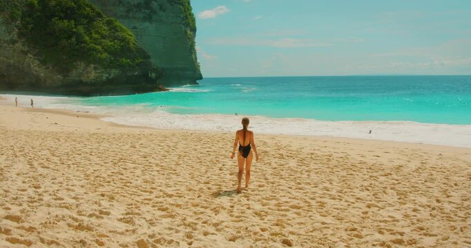 Woman walking toward turquoise ocean on sunny day at Kelingking Beach Bali Nusa Penida