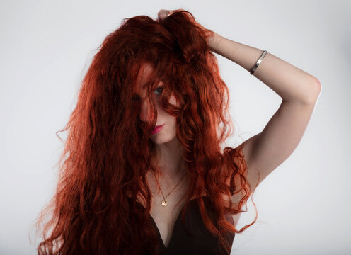 Portrait of an expressive young woman with vibrant curly red hair, hands in hair posing playfully with a kissy face, white background.  Fashion, beauty, lifestyle, and advertising.