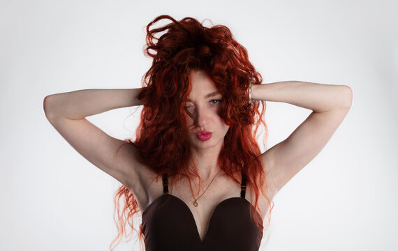 Portrait of an expressive young woman with vibrant curly red hair, hands in hair posing playfully with a kissy face,  white background. Ideal for fashion, beauty, lifestyle, and advertising.
