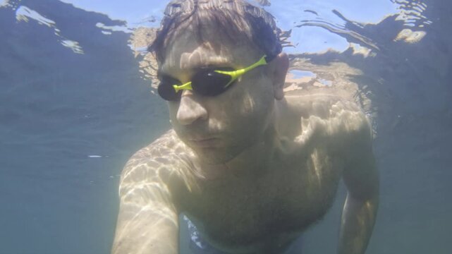 Man holding breath underwater with swimming goggles. Sunlight ripples creating patterns on skin. Summer leisure and aquatic exercise