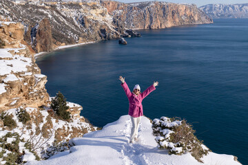 Young woman wearing pink winter coat stands with arms raised on snowy cliff overlooking a calm blue sea during sunny day © svetograph