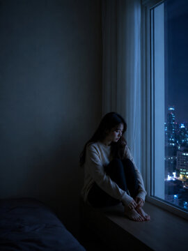 Lonely woman sitting by window at night, city lights outside, feeling sadness and solitude
