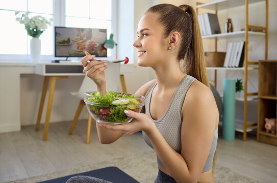 Woman eating salad after home workout fitness. She smiles in sportswear holding a fresh bowl. Light home setting aids yoga, wellness and nutrition themes. Clear and healthy lifestyle concept
