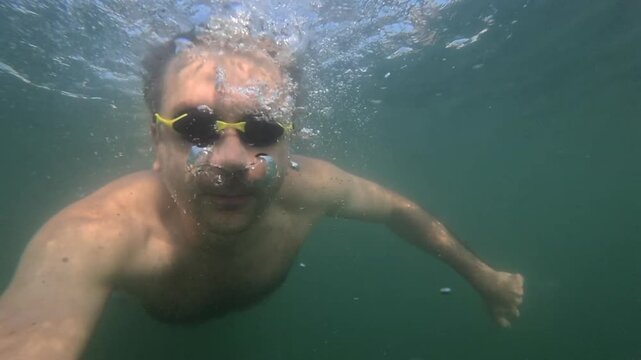 Man wearing swimming goggles exploring underwater, exhaling air bubbles. Recreation, sport and leisure concept