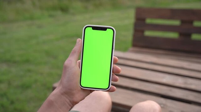 Hand holds phone with green screen outdoors on wooden bench. Person relaxed, seated, focused on device. Perfect for app demos or advertising mockups. Natural daylight, grassy background
