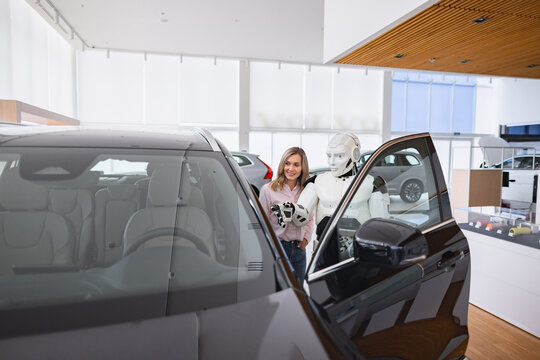 A humanoid robot assists a woman by opening a car door in a modern dealership showroom