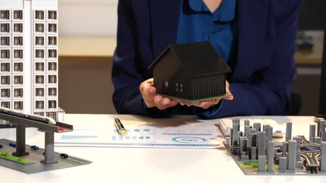 Woman holding a small house model over a desk with architectural plans and city building models, showing real estate planning and property development