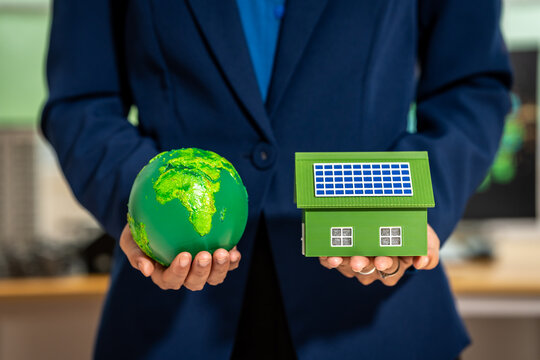 Business person holding a green globe and a small house with solar panels, symbolizing sustainable living, renewable energy, and eco friendly investment.