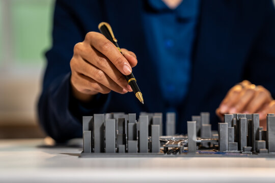 Close-up of a business person holding a pen over a small building model on a table, showing planning, design, and project review
