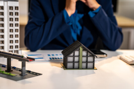 Small model house on a desk with a blurred business person in the background, showing real estate planning, property investment, and home buying concept.