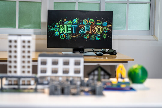 Office desk with a monitor showing net zero and sustainability icons, with blurred building models in the foreground