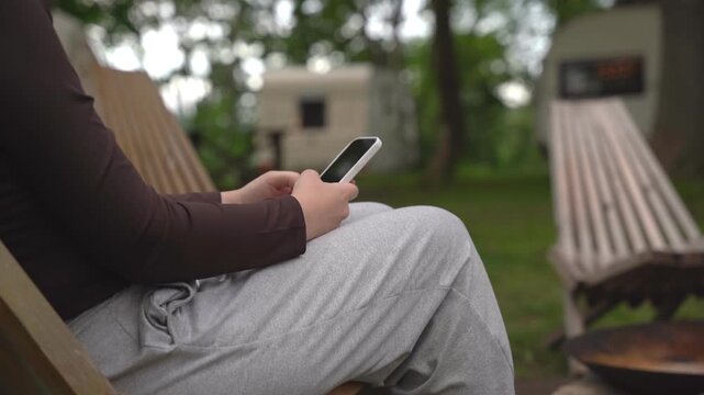 Person sits on wooden chair, focused on phone. Relaxing outdoors near campfire pit. Calm, peaceful atmosphere with green grass and trees. Ideal for travel, camping, or digital detox themes
