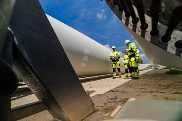 Fototapeta premium Four technicians in yellow and navy coveralls stand in a loose group under the bolted root end of a turbine blade that is parked on its cradle in a component laydown yard