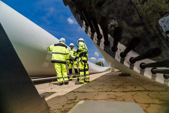 A maintenance crew clusters at the root end of a stored wind turbine blade, one member carrying a black equipment case as the group studies the bolted flange above their heads
