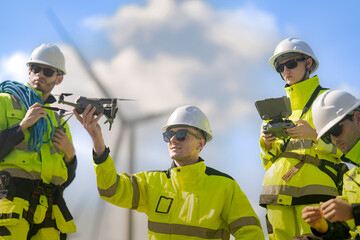 Fototapeta premium Three wind farm technicians press close together as one holds a small drone on his palm and another cups the controller screen, working in bright sunshine under the rotors