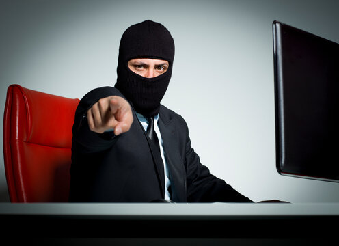 Masked businessman in suit pointing finger at camera at desk cyber threat and extortion concept isolated on white background.