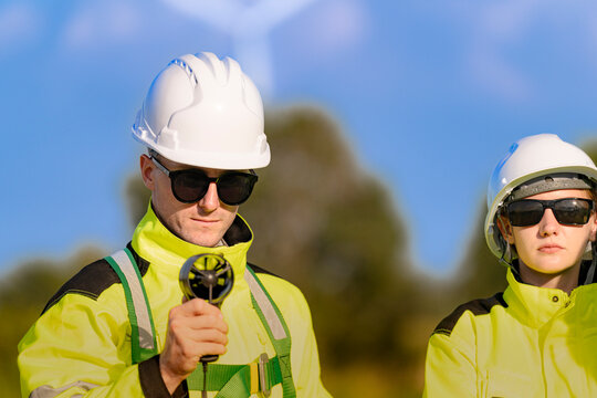 Portrait of a clean-shaven wind technician aiming a handheld vane anemometer toward the camera, with a female colleague standing just out of focus at his right shoulder