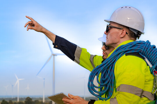 A wind technician throws an arm out to the right to indicate a distant turbine while his colleague watches from behind, the two pressed close in the tight vertical space