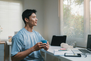 Fototapeta premium Asian man smiling, drinking coffee and looking out window, taking a break from working on laptop in home office