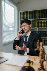 Young male lawyer discussing legal matters on a smartphone, holding a pen, with law books and...