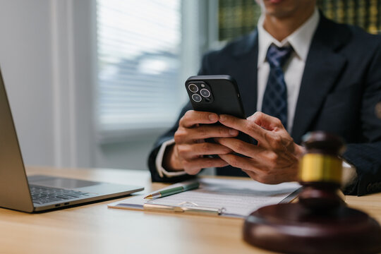 Professional lawyer holding and using a smartphone at an office desk with a gavel, laptop, and legal documents