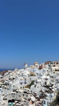 Santorini island, Greece. White cycladic architecture and famous windmills in Oia town. Blue sea and the blue sky at sunny day. Panoramic view. Travel and summer vacation concept. Vertical video
