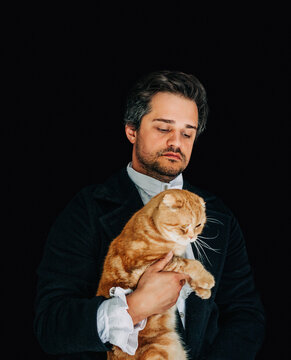 19th century European nobleman in tailcoat holding a red Scottish Fold cat, black background.