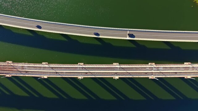 A car drives on the top bridge while shadows of both bridges fall on the green water below, showing a unique perspective of the scene during daylight.