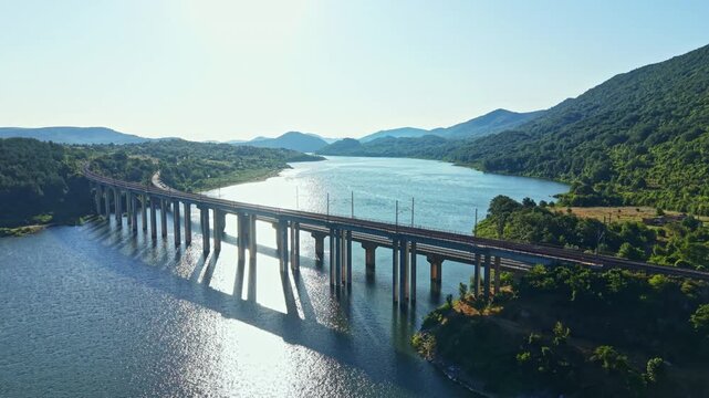 A bridge connects two land areas over a body of water. The sun shines down on the scene, and mountains rise in the distance. The railway runs along the bridge structure.