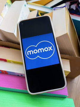 Braunschweig, Germany - August 18, 2026: Cell phone with blue momox platform application icon lying on top of stacked literature, sustainable lifestyle concept of recycling, second hand marketplace.