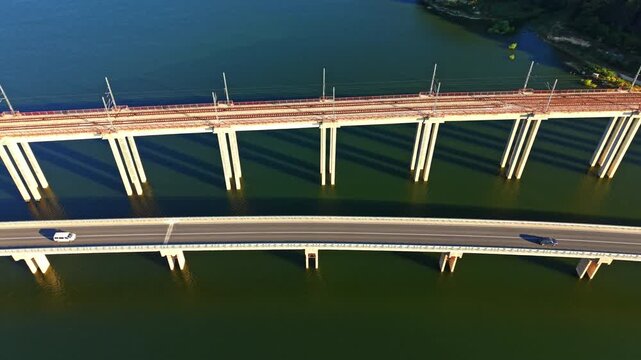 A bridge spans over water with train tracks positioned on top. Below the bridge, a road runs alongside the water. The scene is captured from an aerial perspective in the morning light.