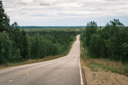 Straight empty asphalt highway dipping through dense taiga pine forest wilderness under cloudy summer skies in Finnish Lapland, Northern Finland.