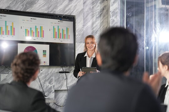 Blonde businesswoman gestures warmly while holding a tablet and presenting colourful charts on a wall screen to seated colleagues in a sleek marble-walled boardroom in Bangkok.