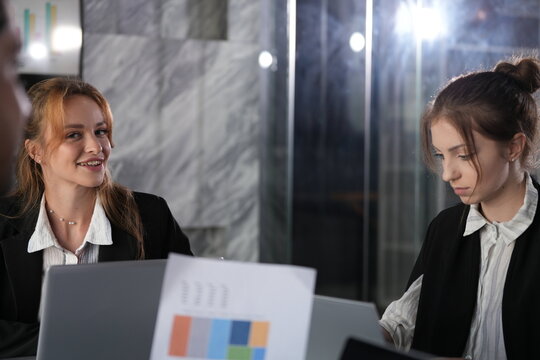 Amused smile with lowered eyes from the ginger haired woman while her colleague in the bun turns a report page during their late night desk session in the coworking office.