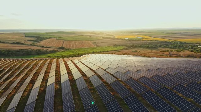 Panels are lined up in rows on a slope, reflecting light as the sun sets in the background. Green fields stretch into the distance, showing a rural landscape.