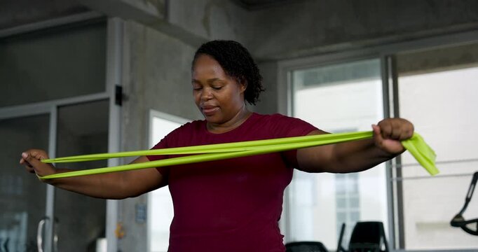 African American plus size woman stretches resistance band across chest, extending arms during shoulder mobility exercise in modern gym. Curvy athlete builds strength, confidence, wellness routine.
