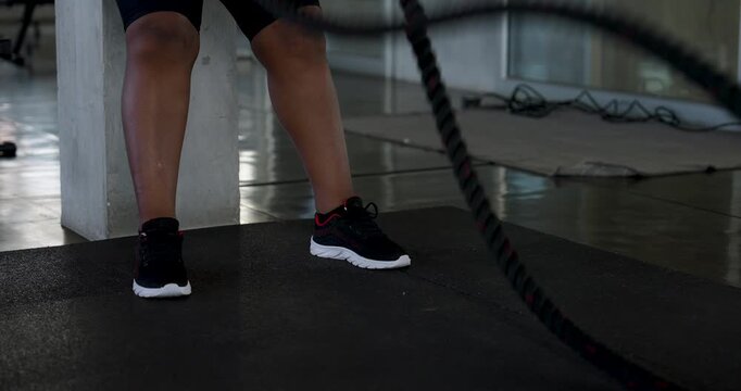 Close view of adult male athlete stepping on gym floor while battle ropes swing during strength training, with African American plus size woman exercising behind. Endurance workout, active lifestyle.