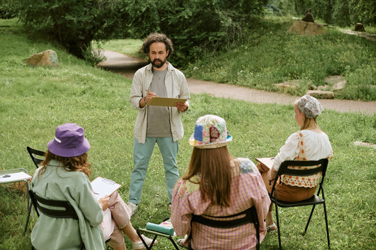 Middle aged Caucasian man teaching outdoor drawing master class to group of young adult women in park, promoting creative education and community art workshops